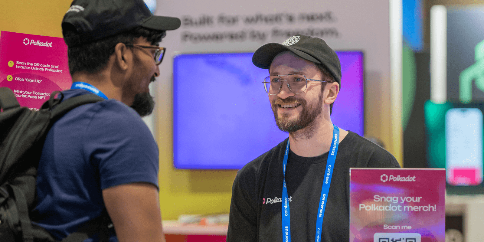 Polkadot team member smiles while talking to a booth visitor at Consensus 2025. Nearby signage shows steps to claim merch and begin onboarding.