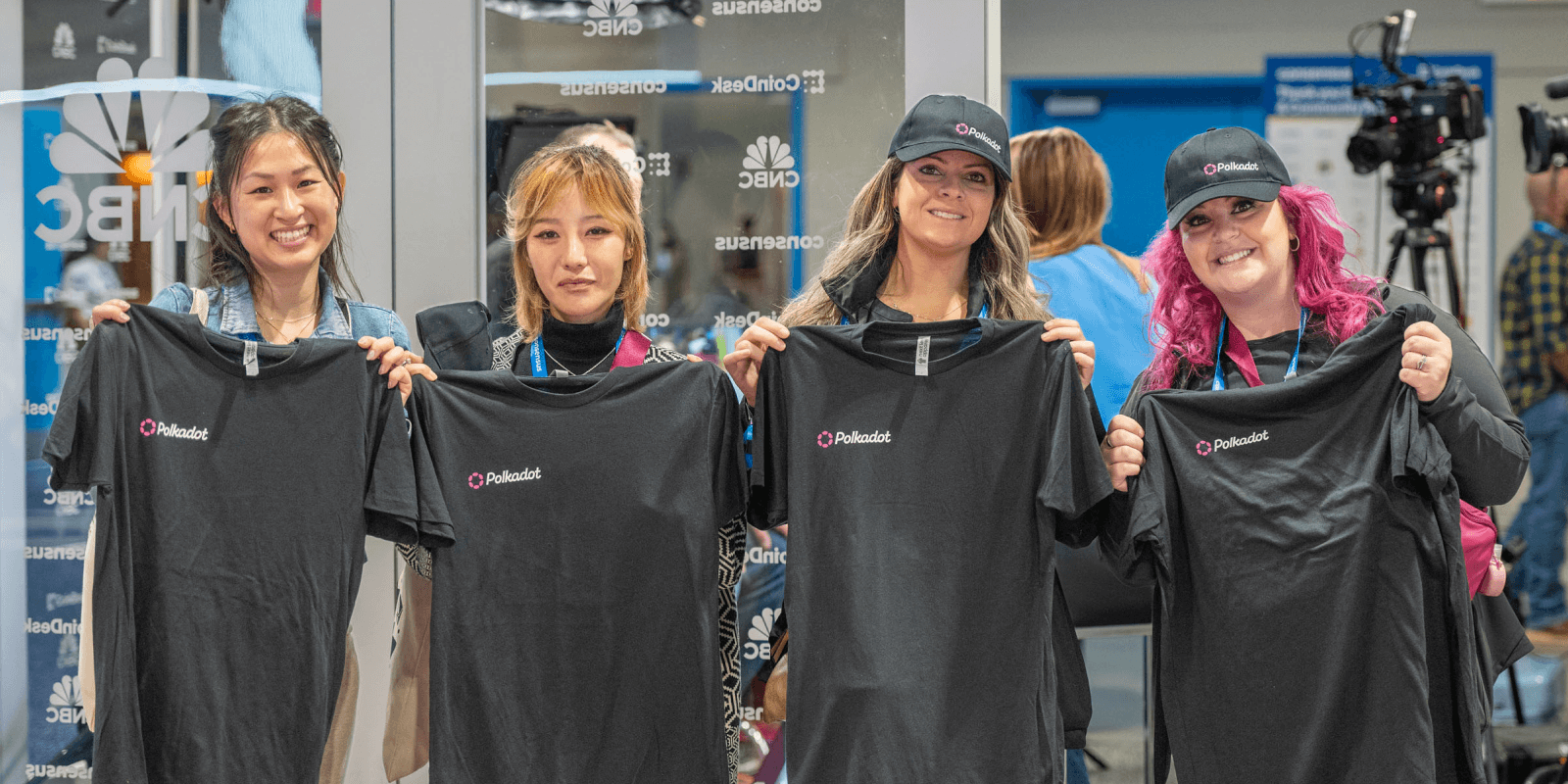 Four attendees at Consensus 2025 smile while holding up Polkadot t-shirts, showing off swag earned through the event's onboarding experience.